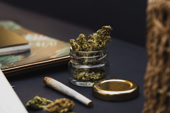 Jar of cannabis buds, gold lid, notebooks, joint on black background