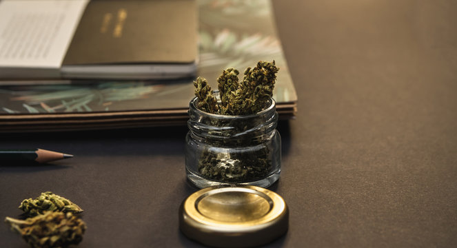 Marijuana buds in open glass jar with gold lid, notebooks and pencil on black background