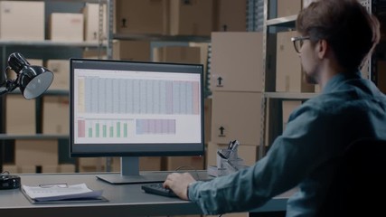 Warehouse Inventory Manager Works with a Spreadsheet on a Personal Computer while Sitting at His Desk. In the Background Shelves Full of Cardboard Box Packages Ready For Shipping. - Powered by Adobe