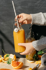 still life, female hands holding orange juice in glass bottles with striped tubules on a wooden table with slices of chopped orange and mint