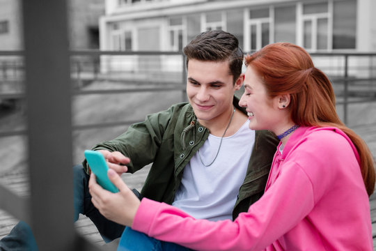 Looking at smartphone. Positive young girl laughing and holding modern smartphone while her curious boyfriend pointing to the screen of it