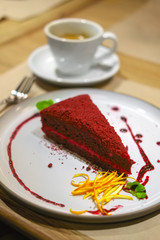 Piece of cherry cake with cranberry syrup decorated with mint and orange zest, cup of coffee on background.