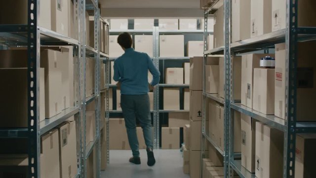 Worker Carrying Cardboard Box Walks Through Warehouse Storeroom with Rows of Shelves Full Cardboard Boxes, Parcels, Packages Ready For Shipment.