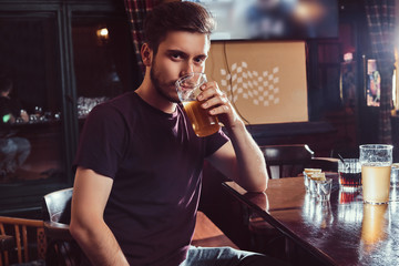 Young happy handsome guy resting in the bar or pub drinks beer at wooden counter.