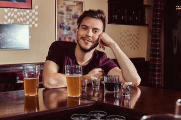 Young happy handsome guy resting in the bar or pub, sitting with glass of beer at wooden counter.