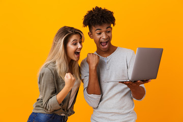 Happy young loving couple standing isolated over yellow background using laptop computer make...