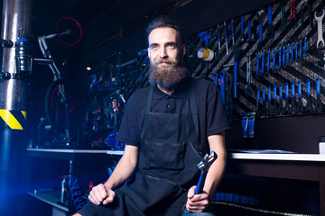 Fototapeta na wymiar Portrait of small business owner of young man with beard. Guy bicycle mechanic workshop worker sitting with tool in his hand in a working black clothes in an apron in the background of a bicycle shop.