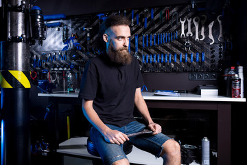 Fototapeta na wymiar Portrait of small business owner of young man with beard. Guy bicycle mechanic workshop worker sitting with tool in his hand in a working black clothes in an apron in the background of a bicycle shop