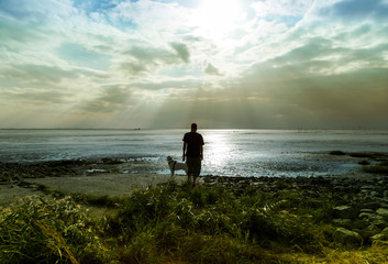 Erholung an der Nordseeküste, Mann mit Hund allein am Wattenmeer bei Bremerhaven im Gegenlicht, Sturmwarnung