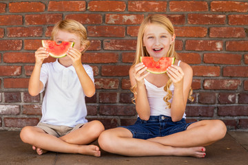 Cute Young Cuacasian Boy and Girl Eating Watermelon Against Brick Wall