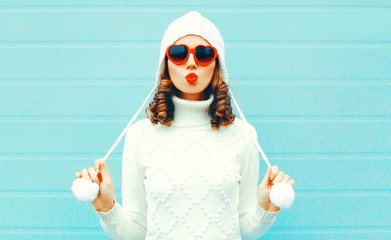 Portrait beautiful woman in white knitted sweater, hat, sends air kiss on blue background