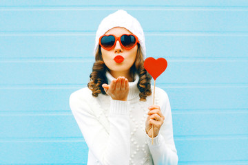 Portrait pretty woman blowing red lips sends air kiss with lollipop on stick heart shape in white knitted hat, sweater on blue background
