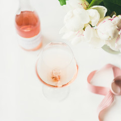 Rose wine in glass and bottle, pink decorative ribbon, peony flowers over white background, copy space, square crop. Summer celebration, wedding greeting card, invitation concept