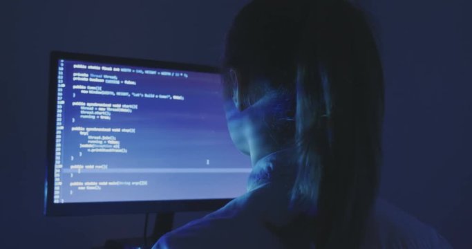 Experienced IT woman programmer works on a new program. Young woman hacker writes the source code of the software at dark room. Back view.