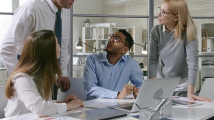 Mid-aged male and female business professionals explaining something on laptop screen to young diverse coworkers while brainstorming in the office