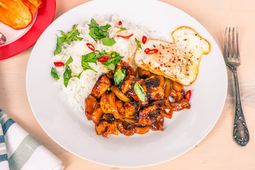 Thai cuisine - chicken thai basil, chicken in sauce with rice and fried egg, on a plate on a light rustic table