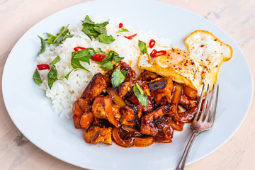 Thai cuisine - chicken thai basil, chicken in sauce with rice and fried egg, on a plate on a light rustic table