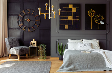 Real photo of dark grey bedroom interior with molding and paintings on walls, double bed with pillows, gold lamp and floral armchair