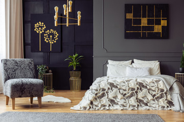 Real photo of floral armchair standing in dark bedroom interior with two paintings, gold lamp and fur coverlet on king-size bed with pillows - obrazy, fototapety, plakaty