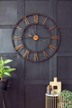 Real photo with close-up of big clock hanging on black wall in dark living room interior with gold accessories and fresh plants