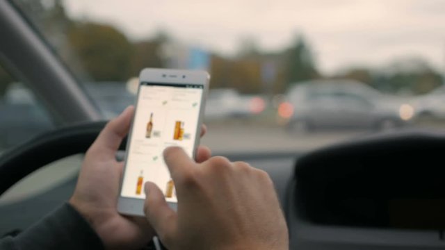 A man looks at the alcoholic  goods in the online grocery store, sitting in the car. Smartphone online shopping. Driver orders whiskey in an online store. Screen is blurred. Focus on finger hand.