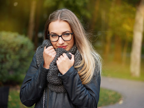 portrait of a beautiful stylish young blonde girl with make-up in black glasses leather jacket and knitted scarf freezes up smiles in the autumn cold park