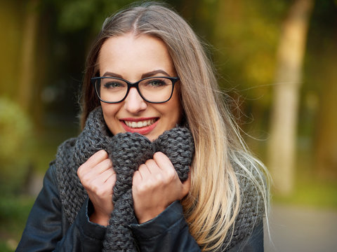 portrait of a beautiful stylish young blonde girl with make-up in black glasses leather jacket and knitted scarf freezes up smiles in the autumn cold park