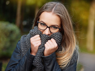 portrait of a beautiful stylish young blonde girl with make-up in black glasses leather jacket and knitted scarf freezes up smiles in the autumn cold park