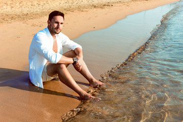 Handsome man in white shirt standing on a beach seaside looking hot on summer day enjoying life. Brutal Male model poses with wet short in water naked chest out, with smirk sitting depressed.