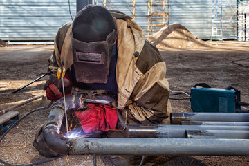 A builder welder in brown working clothes welds a metal product with arc welding machine in the factory, a lots of tools and constraction, bright lights and sparks