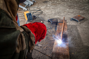 Close up of a builder welder in brown working clothes welds a metal product with arc welding machine in the factory, a lots of tools and constraction, bright lights and sparks