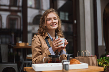 Young joyful woman in trench coat happily looking in camera holding white cup of tea in hands while spending time outdoor at cozy cafe terrace