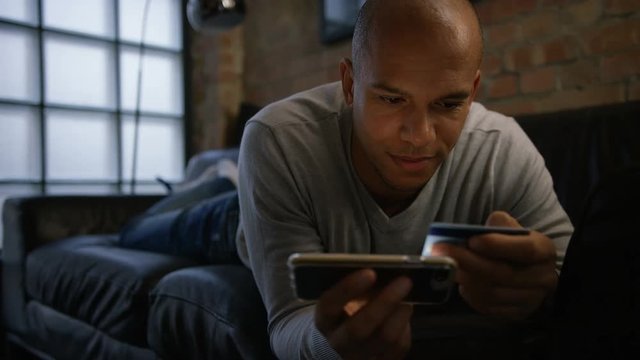 Young male on his couch using his phone and credit card for online shopping