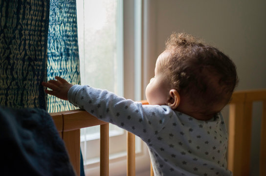 Baby boy standing up in crib looking out through window