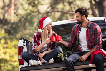 young couple in santa hats clinking with red cups while sitting on pickup truck with christmas tree - Powered by Adobe