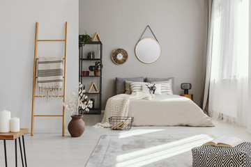 Mirror and clock above bed in bright bedroom interior with pouf and flowers next to ladder. Real...