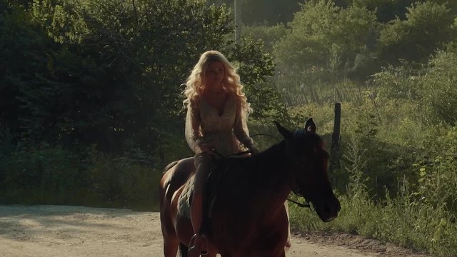 Beautiful blonde girl is riding horse in summer day in a forest. She is walking out from woodland on sunny road in daytime