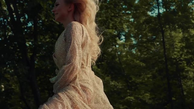 Alluring girl with blonde curly hair is riding a horse. She is galloping between trees in a woodland in sunny day, moving along camera