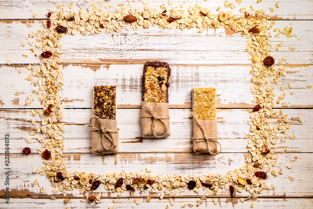 Wall mural Various granola chocolate energy bars in row with scattered mixed nuts, cereals & dried fruit, grunged white wood table background. Healthy vegan fitness food snack. Top view, copy space, close up.