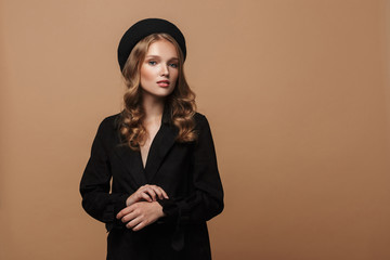 Young beautiful woman with wavy hair in black coat and beret dreamily looking in camera over beige...