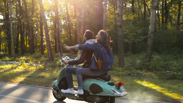 Young couple riding on their scooter at the road in the forest. Woman shooting video on the action camera, using selfiestick.