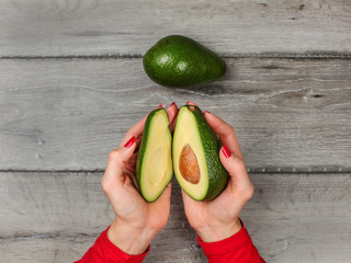 Tabletop view, young woman hands holding two halves of avocado cut in half, over gray wood desk,...
