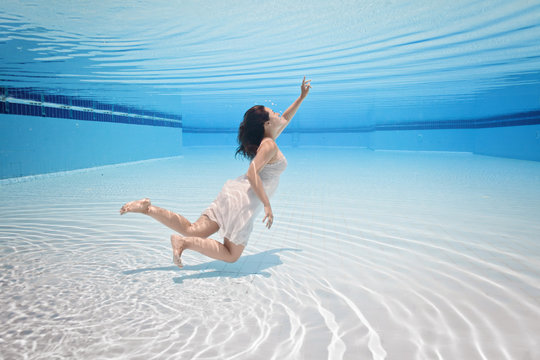 Young Attractive Girl Swim Underwater In Swimming Pool In Pink Dress. Diving And Floating Under Water Surface. Playing With Reflections And Waves. Touch Water Mirror With Her Hand