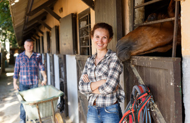 Mature positive female farmer standing near horse at stable