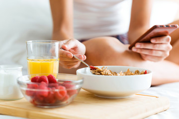 Young woman having breakfast and texting with her smartphone in the bedroom.