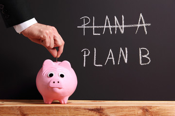 Man in suit dropping a coin into a piggy bank located in front of a blackboard where one can read: Plan A and Plan B.  Plan B has been crossed out