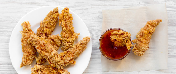 Spicy chicken strips on white plate with sauce over white wooden surface, overhead view. Flat lay,...