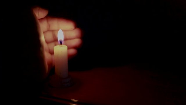 Candle burning and almost is blown out by the wind but is then blocked by a hand so it won't blow out. The room is completely dark except for the candles light on the table and the hand.