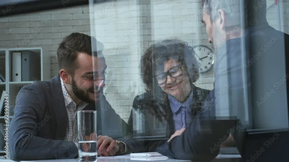 Poster medium shot of three colleagues viewing something on tablet computer and smiling when sitting at des