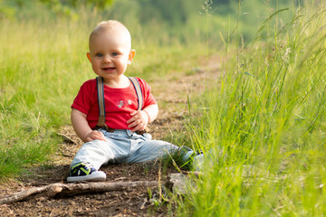 Naklejka na ściany i meble Little baby boy sitting on a forest path on a nice sunny day. A blond newborn with blue eyes is wearing bib shorts.